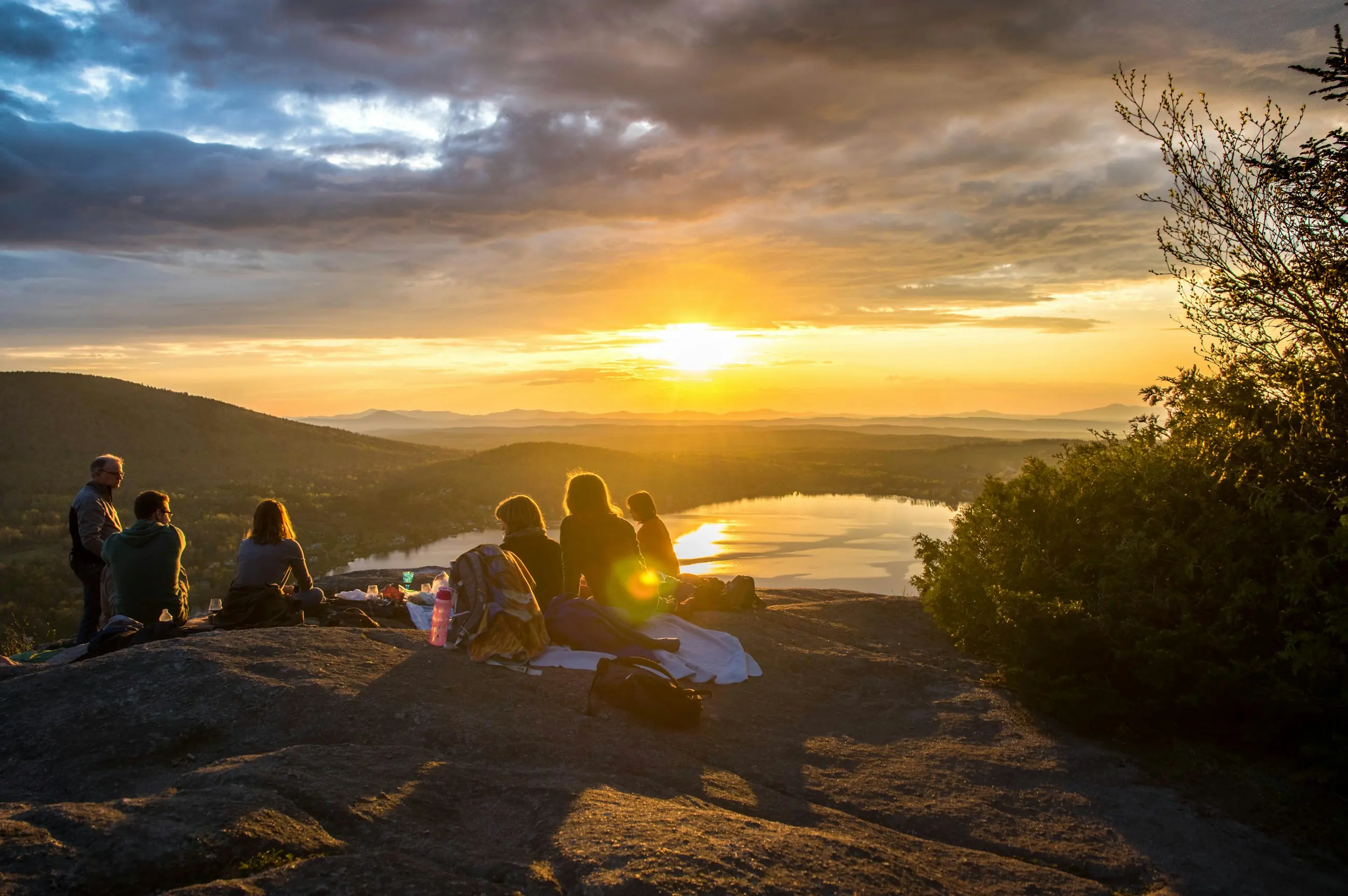 Amigos sentados no alto de um monte, olhando o nascer do sol sobre um lago